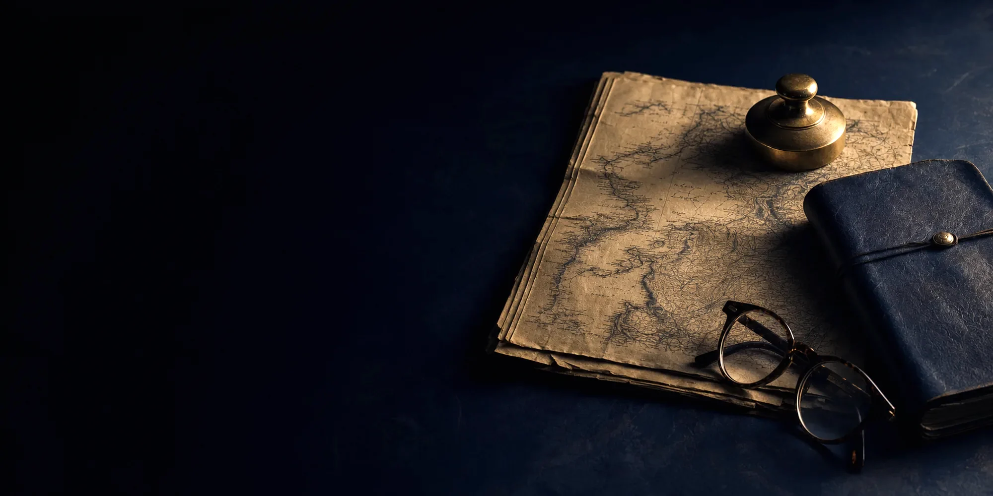 An archival map, a notebook and reading glasses lit low on a dark desk