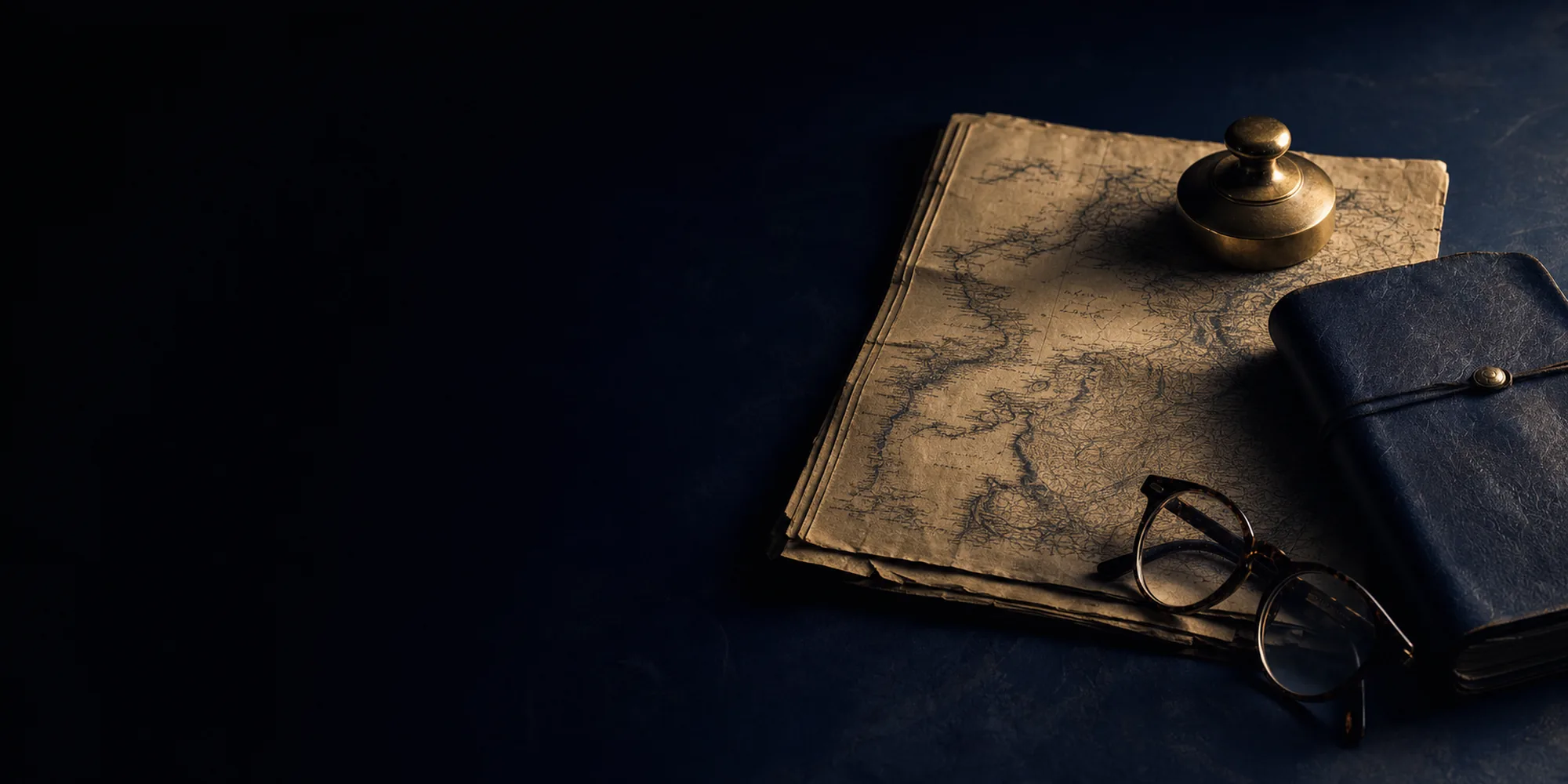A leather notebook, inkwell and reading glasses on a dark desk