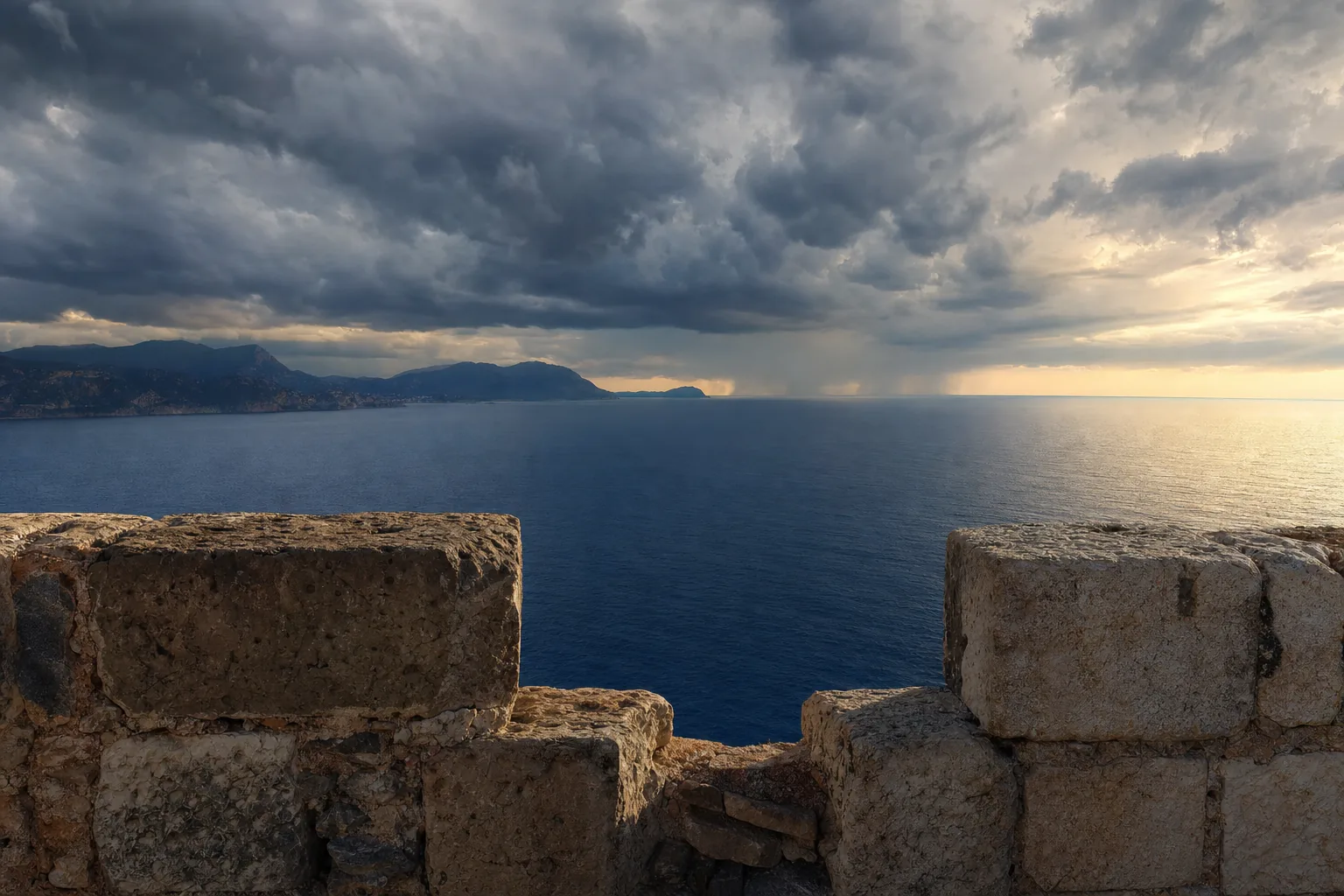 The open sea and a distant headland seen over a stone parapet at dusk